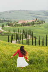 Young happy woman in red sweater and white skirt on background on spring Tuscany landscape. Dancing and enjoying view of cypress alley and yellow flowers field