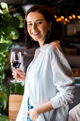 cheerful woman standing with hand in pocket while holding glass with red wine