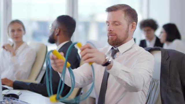 Lockdown of businesspeople sitting at desktops, working while Caucasian businessman starting warming up his arms using chest expander while sitting on office chair and then continuing typing message