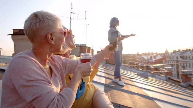 Medium shot of two smiling middle-aged women sitting together on roof, making photos using smartphone while grey-haired woman standing nearby with glass of wine in her hands