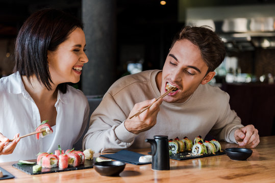 selective focus of man holding chopsticks while eating sushi near happy woman in sushi bar