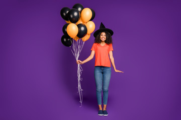 Full length body size photo of cheerful positive cute nice charming pretty girlfriend wearing jeans denim witch hat orange t-shirt holding air balls smiling toothily isolated bright violet color