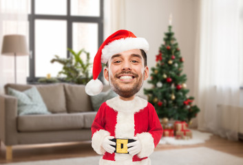 emotion, expression and winter holiday concept - happy smiling young man in santa claus costume over christmas tree on home background (funny cartoon style character with big head)