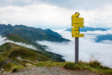 Yellow signposts pointing in different directions on the top of a mountain in the Austrian Alps with a cloudy valley and a beautiful panorama in the background