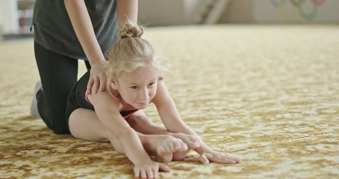 Crop coach helping girl with stretching exercise, performing stretching exercise with straight and split legs on carpet in gym