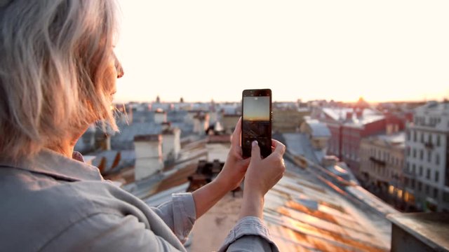 Back view of grey-haired middle-aged woman standing on roof in historical center of city with cellphone in her hands and making photos