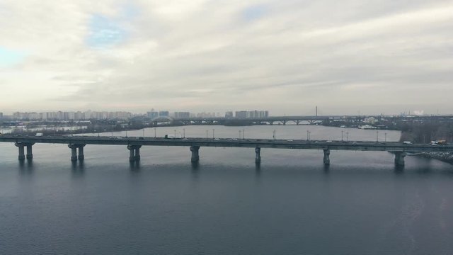 Traffic on a bridge over a wide river in the winter. Drone is approaching the automobile bridge, view from above. Cars move on the bridge in both directions. Cold tinting in the winter.