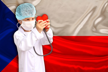child, boy, in a white doctor’s coat, hat and mask attached a stethoscope to a red heart model,...