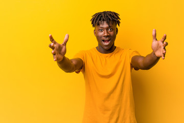 Young black man wearing rastas over yellow background feels confident giving a hug to the camera.