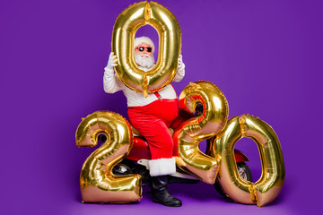 Full body photo of fat santa man sitting on bike holding big air newyear 2020 numbers balloons congratulating people wear sun specs x-mas costume isolated purple background