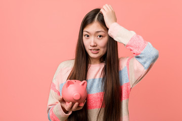 Obraz na płótnie Canvas Young asian woman holding a piggy bank being shocked, she has remembered important meeting.