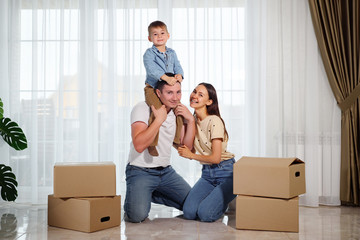 happy smiling family against the background of panoramic windows. Son sits on dad's shoulders near beautiful mother among cardboard boxes.