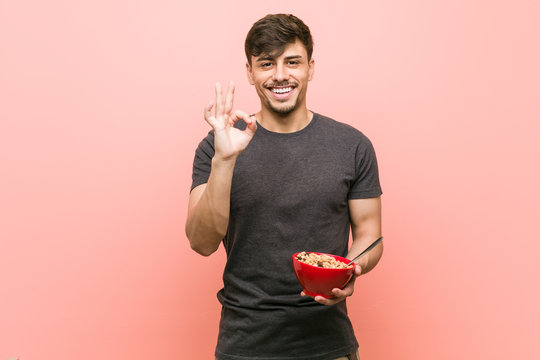 Young hispanic man holding a cereal bowl cheerful and confident showing ok gesture.