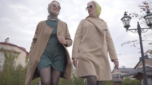 Bottom view of two mature Caucasian women wearing beige coats, headscarves and sunglasses walking in the city park. Elegant senior female friends travelling together.