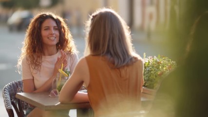 Slowmo medium shot of happy young woman with curly hair and her female friend sitting at table in outdoor seating area of cafe and chatting on sunny summer day