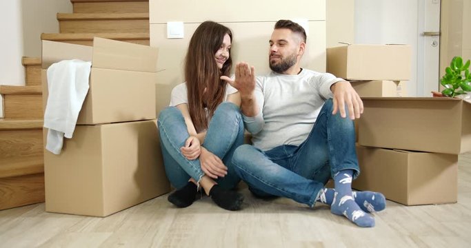Happy man and lady sitting on floor in new apartment among cardboard boxes, giving high-five, moving to new flat