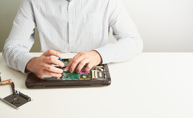 Man repairs computer. A service engineer in shirt repairs laptop, at white Desk against white wall.