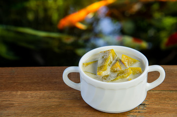 Yummy pumpkin in sweet coconut milk in white bowl, popular Thai dessert on wooden table with blurred fancy carp fish in pond background