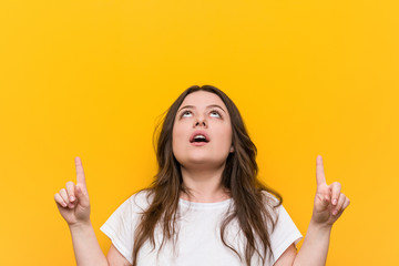 Young curvy plus size woman pointing upside with opened mouth.