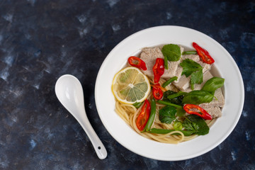 Asian soup with wheat noodles, slices of meat, red chili peppers. Decorated with mint leaves, basil, green onion feathers. There are carrots and broccoli. A healthy, dietary product. Copy space.  