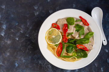 Asian soup with wheat noodles, slices of meat, red chili peppers. Decorated with mint leaves, basil, green onion feathers. There are carrots and broccoli. A healthy, dietary product. Copy space.  