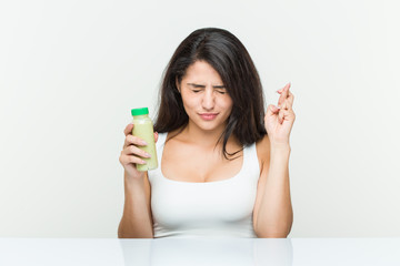 Young hispanic woman holding a vegetable drink crossing fingers for having luck