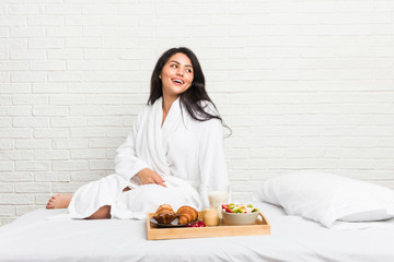 Young curvy woman taking a breakfast on the bed looks aside smiling, cheerful and pleasant.