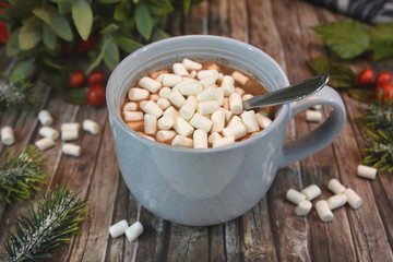 Gray mug with chocolate drink topped with small white marshmallows and chocolate and cinnamon powder on wooden desk with winter decor arrangement