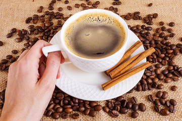 White cup with hot americano and cinnamon on a saucer. Coffee beans on burlap. Hand holds a cup. The concept of making coffee. Flat lay, top view, minimalism.