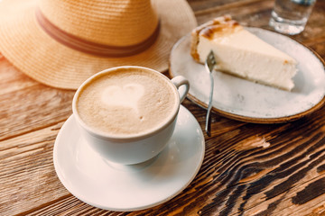 A Cup of cappuccino with foam in the shape of a heart and hat with a piece of cheesecake on a vintage wooden background in a cafe