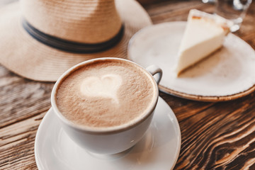 A Cup of cappuccino with foam in the shape of a heart and hat with a piece of cheesecake on a vintage wooden background in a cafe