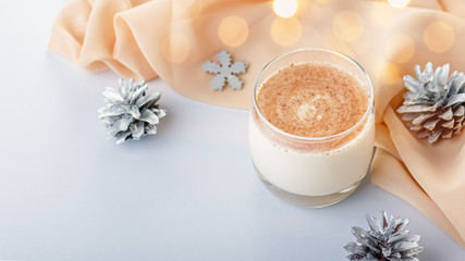 Traditional Christmas cocktail Eggnog with eggs, alcohol, grated nutmeg and cinnamon closeup. Sweet traditional drink on grey table with beige decorations, burning lanterns and pine cones