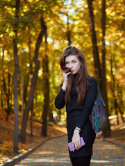 Natural lifestyle outdoors portrait of young adorable redhead woman in black dress with trendy shopper bag emotionally posing in autumn city park with gorgeous golden fall leaves background