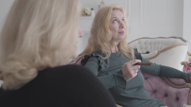 Smiling Mature Caucasian Women Sitting In Comfortable Armchairs Holding Money And Cigars And Talking. Old Female Friends Gossiping About Their Life.