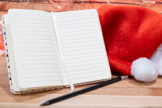 notebook,red Christmas hat and pencil on wooden table on background christmas garland