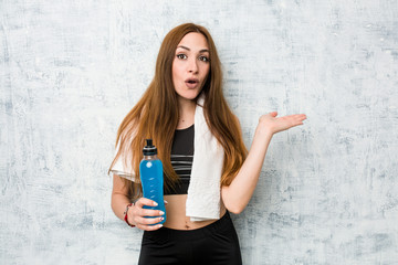 Young caucasian sportswoman holding a isotonic drink impressed holding copy space on palm.