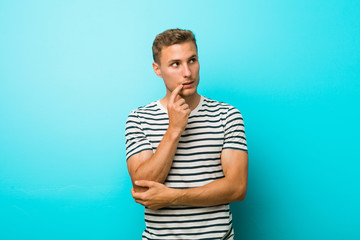 Young caucasian man against a blue wall relaxed thinking about something looking at a copy space.