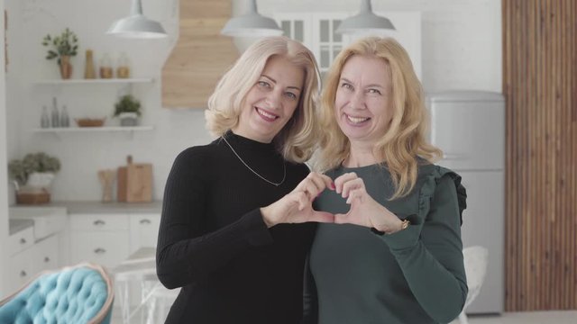 Smiling mature Caucasian women standing in luxurious kitchen and falding fingers into heart shape. Rich senior female friends spending free time together.