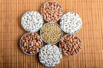 Seven bowls with organic fresh green lentil beans, red and white beans on a wooden table displayed as a flower in warm light, top view
