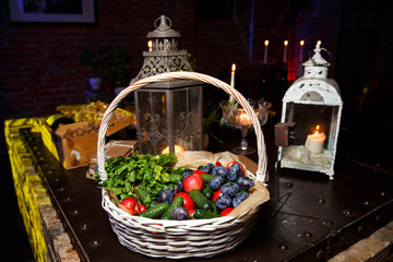 Basket with parsley, cucumbers, tomatoes, apples, plums on background of lantern with candles.