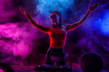 Young sexy woman dj enjoys playing music, raised her hands up. Headphones and a DJ mixer on the table. Colored smoke on the background - obrazy, fototapety, plakaty