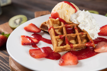 The strawberry waffle, topping with whipped cream, vanila ice cream and strawberry sauce and fresh on a white plate. Decorated with fresh fruit. Dark brown real wood chopping and background.