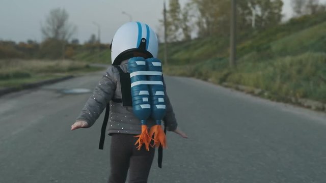 Little girl wearing white helmet, jetpack and running on rural road in slow motion. Tracking of kid pretending to be pilot