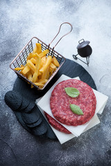 Raw burger cutlets made of marbled beef meat, french fries and black potato chips, high angle view on a grey stone background