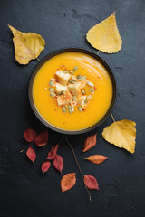Pumpkin cream-soup over black stone background with autumn leaves, top view, vertical shot
