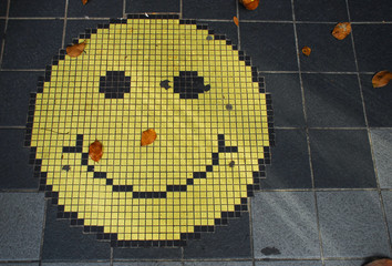 Mosaic of an smiling face emoticon on a gray tiles sidewalk, and some fallen tree leaves