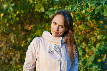 Fototapeta premium Smiling long-haired beautiful young woman wearing light pink coat looking at camera. Blurry leaves background.
