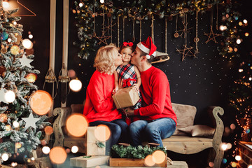 Christmas family with baby smiling and kissing near the Xmas tree. Living room decorated by Christmas tree and present gift box, the light give cozy atmosphere. New Year theme