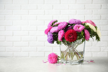 Glass vase with beautiful aster flowers on table against white brick wall. Space for text