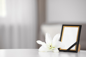 Obraz na płótnie Canvas Funeral photo frame with black ribbon and lily on white table indoors. Space for design